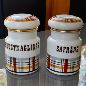 Op de afbeelding: Twee witte keramische kruidenpotten met gouden deksels en geruite ontwerpen. De potten zijn gelabeld met "KRUSTNAGLIŅAS" en "SAFRĀNS".