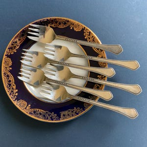 Puede incluir: Seis tenedores plateados colocados sobre un plato decorativo. El plato tiene un centro blanco, un borde azul marino y diseños dorados. Los tenedores están colocados en diagonal, con sus púas apuntando hacia afuera. Vajilla clásica.
