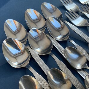 May include: A collection of shiny, silver spoons and forks arranged on a dark blue surface. The spoons have a rounded bowl and a handle with a decorative pattern. The forks have three tines and match the spoon's handle design. The silverware reflects the light.