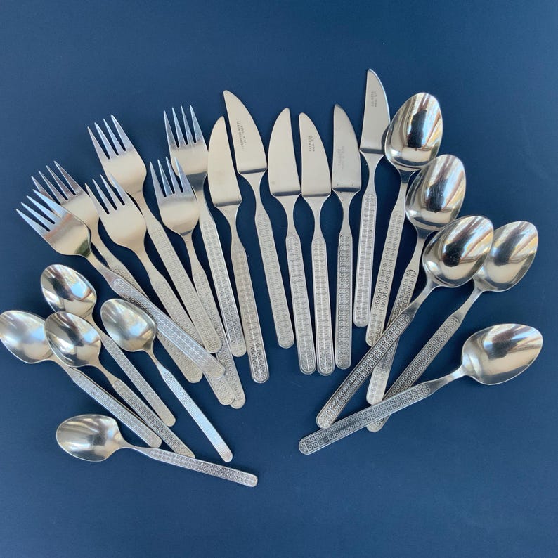 May include: A collection of stainless steel silverware, including forks, knives, and spoons, arranged on a dark blue surface. The silverware has a textured pattern on the handles and a shiny, reflective finish.