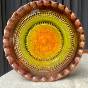 May include: A round ceramic pie dish with a ruffled edge. The dish features a gradient color scheme transitioning from a dark brown rim to a bright lime green middle and a warm yellow center. An etched star pattern is centered on the base.