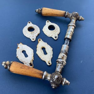 May include: Antique door hardware set featuring two door handles with wooden grips and ornate metal detailing, along with four white, decorative keyhole covers. The hardware has a vintage, weathered appearance.