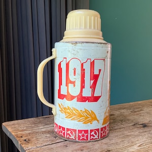 May include: A white metal thermos with a red and yellow design. The thermos has a handle and a lid. The thermos has the year 1917 printed on the front. The thermos has a red star and hammer and sickle symbol on the bottom.