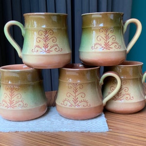May include: Set of five brown ceramic mugs with a green glaze and a brown floral design. The mugs have a rounded shape and a handle.