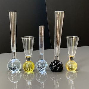 May include: Five glass bud vases with round bases. The bases are clear glass with air bubbles, yellow glass with air bubbles, clear glass with air bubbles, black glass, and yellow glass with air bubbles.