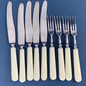 Peut inclure: Un ensemble de couverts anciens, comprenant des couteaux et des fourchettes, disposés sur un fond bleu. Les couteaux ont des lames argentées et des manches blanc cassé. Les fourchettes présentent un design similaire, avec des dents argentées et des manches blancs cassé.