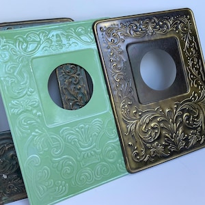 Puede incluir: Primer plano de varias cubiertas decorativas de interruptores de luz. Una es de color verde claro y otra de color bronce. Ambas tienen diseños florales en relieve y una abertura cuadrada para el interruptor.