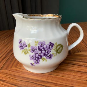 Peut inclure: Une crémier en porcelaine blanche avec un bord doré et un motif floral avec des fleurs violettes et des feuilles vertes.