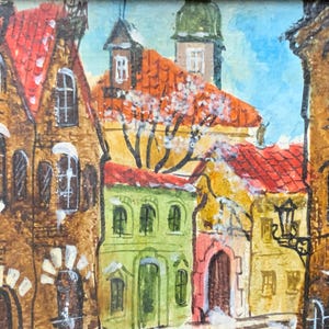 Op de afbeelding: Een aquarel schilderij van een Europees dorpstraatje met kleurrijke gebouwen, een boom en een lantaarnpaal. De gebouwen zijn geschilderd in tinten bruin, rood en groen, en de boom is geschilderd in tinten groen en bruin. De lantaarnpaal is geschilderd in zwart en wit.