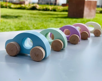 May include: A set of five wooden toy cars in pastel colors. The cars are arranged in a line from largest to smallest, with the largest car being light blue, followed by light green, lavender, pink, and white.