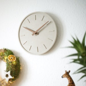 Auf dem Foto sieht man eine Wanduhr aus Beton mit Zeigern aus Walnussholz. Die Zeigermarkierungen sind schlicht mit angedeuteten Strichen. Die Holzzeiger elegant mit einer Anbringung aus Messing. Die Betonuhr ist handgefertigt in Deutschland.