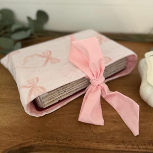 May include: A pink fabric-covered book with a pink ribbon tied around it. The book has a pattern of pink bows on it.
