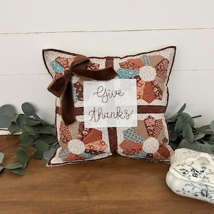 May include: A square quilted throw pillow featuring a patchwork fan pattern in shades of brown, teal, and cream. A white fabric square in the center displays the embroidered text Give thanks in brown thread, accented by a dark brown velvet ribbon bow.