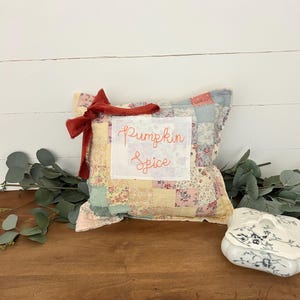 May include: A square patchwork pillow featuring a variety of pastel floral fabric squares in shades of pink, yellow, and blue. A white rectangular patch in the center displays the embroidered orange text Pumpkin Spice, accented by a dark red fabric bow.