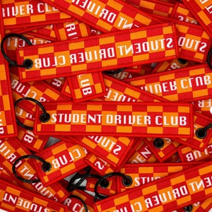 May include: A close-up of a pile of red and orange keychains with the text "STUDENT DRIVER CLUB" printed on them. The keychains are made of a checkered fabric and have a metal ring attached.