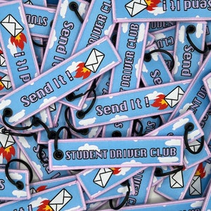 May include: A pile of blue and white keychains with the text "Send It!" and "Student Driver Club". The keychains feature a cartoon image of an envelope with flames.