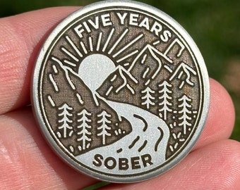 May include: A round silver-toned metal sobriety coin featuring an engraved landscape design. The image depicts a sun rising over mountains, a winding river, and pine trees. The text FIVE YEARS is arched at the top and SOBER is at the bottom.