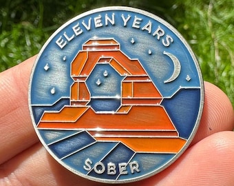 May include: A silver-colored round pin with a blue background and a red and orange image of Delicate Arch in Arches National Park. The pin says "Eleven Years Sober" around the edge.