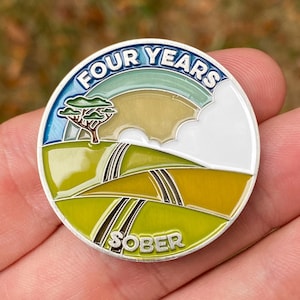 May include: A circular, silver-coloured metal coin with a vibrant enamel design. The coin displays the words "FOUR YEARS" and "SOBER" in white text. The design includes a landscape with a tree, rolling hills, and a rainbow.