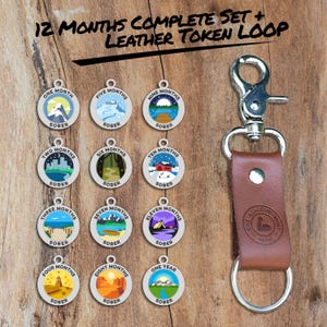 May include: A set of 12 silver-toned metal tokens with colorful illustrations of different landscapes and the words "One Month Sober" to "One Year Sober". The tokens are attached to a brown leather keychain with a silver metal clasp.