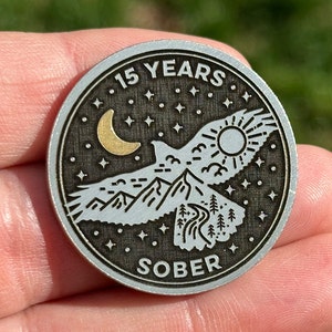 May include: A silver metal coin with a black background and a gold design. The coin features a bird flying over mountains with a sun and moon in the sky. The text "15 YEARS SOBER" is engraved on the coin.