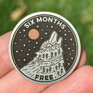 May include: A silver metal token with a black background and a wolf howling at the moon. The token says "Six Months Free" and has a design of a wolf howling at the moon with a mountain range and trees in the background.