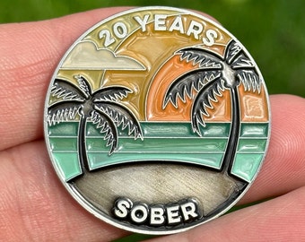 May include: A silver-toned round coin with a sunset scene and two palm trees. The coin has the text "20 Years Sober" on it.