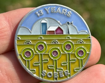 May include: A silver metal pin with a blue and white sky, a white farmhouse with a red roof, and a field of green and yellow sunflowers. The pin says "13 Years Sober".