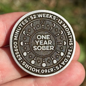 May include: A silver-coloured round coin with the words "ONE YEAR SOBER" in the centre. The coin has an outer ring with the words "52 WEEKS", "12 MONTHS", "365 DAYS", and "8760 HOURS". The coin is held in a hand.