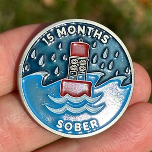 May include: A silver-colored round pin with a blue and white ocean scene. The pin features a red lighthouse with a white top and a white wave. The text "15 MONTHS SOBER" is printed on the pin.