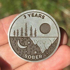 Puede incluir: Una moneda redonda de color plateado con las palabras "3 YEARS" en la parte superior. La moneda presenta un diseño de paisaje con árboles, un sol, una luna, estrellas y la palabra "SOBER".