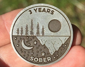 May include: A round, silver-colored coin with the words "3 YEARS" at the top. The coin features a landscape design with trees, a sun, a moon, stars, and the word "SOBER".