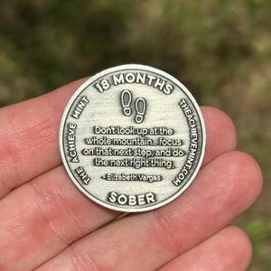 May include: Silver coin with the text "18 Months The Achieve Mint.com" and the quote "Don't look up at the whole mountain... focus on that next step, and do the next right thing. - Elizabeth Vargas" and the word "SOBER".