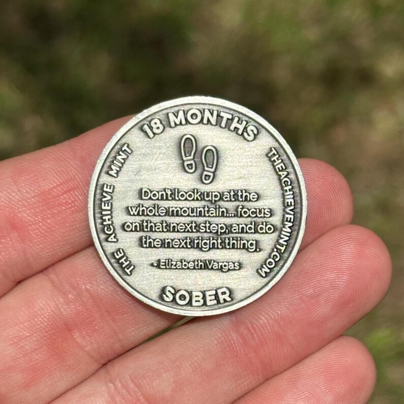 May include: Silver coin with the text "18 Months The Achieve Mint.com" and the quote "Don't look up at the whole mountain... focus on that next step, and do the next right thing. - Elizabeth Vargas" and the word "SOBER".