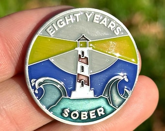 May include: A silver coin with a blue, green, and yellow enamel design. The coin features a lighthouse with a beam of light shining out. The text "EIGHT YEARS SOBER" is printed on the coin.