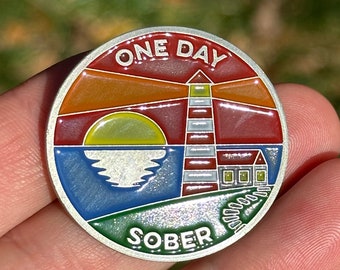 Peut inclure: Une pièce ronde argentée avec un motif en émail coloré. La pièce représente un phare avec un design rayé rouge, blanc et bleu, un soleil jaune, une mer bleue et de l'herbe verte. Le texte "ONE DAY SOBER" est imprimé sur la pièce.