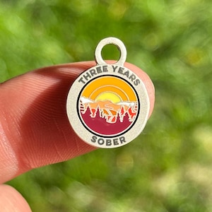 Peut inclure: Breloque en argent avec une scène de coucher de soleil et le texte "THREE YEARS SOBER" en cercle.