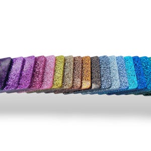 Peut inclure: Une collection d'objets en forme de losange recouverts de paillettes dans un spectre de couleurs, allant du violet foncé au turquoise et vert vif. Les objets sont disposés en rangée, présentant une variété de teintes chatoyantes.