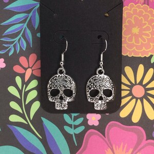 May include: Silver dangle earrings with a detailed sugar skull design. The earrings are displayed on a black card with a floral background.