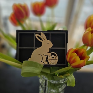 Puede incluir: Decoración de Pascua con un conejito marrón claro sosteniendo una cesta de huevos, colocado frente a una rejilla negra. El conejito se coloca frente a un jarrón de tulipanes con pétalos rojos y amarillos, creando un ambiente primaveral.
