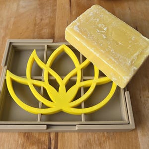 Puede incluir: Un jabonera amarilla con un diseño floral y una pastilla rectangular de jabón amarillo. La jabonera es de color beige claro y tiene un patrón de rejilla. El jabón está sobre la jabonera.