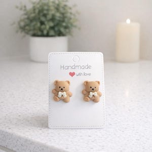 May include: A pair of teddy bear stud earrings on a white card. The earrings are light brown with white bow ties. The card reads "Handmade with love" in black text with a red heart. The background includes a plant and a lit candle.