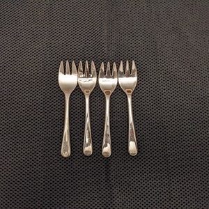 May include: Four silver forks with a polished finish are arranged in a row on a black textured surface.