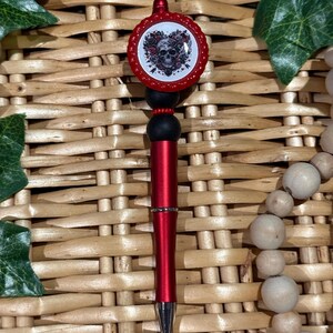 May include: Red pen with a black and red skull and flower design on the top. The pen is decorated with black and red beads.