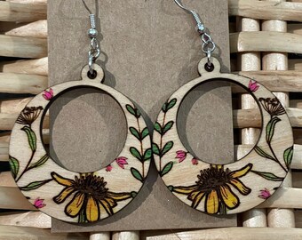 May include: A pair of wooden earrings with a floral design. The earrings are round and have a silver hook. The flowers are yellow, pink, and green.
