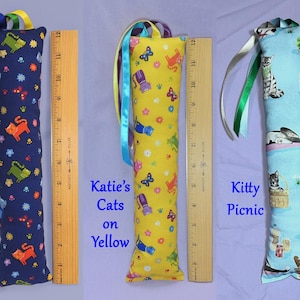 May include: Three cat toy pillows, each approximately 30 cm long, with colourful patterns and ribbons. One is navy blue with orange and green cats, one is yellow with cat and butterfly designs, and the last is light blue with cat illustrations. Text on the pillows reads "Katie's Cats Blue", "Katie's Cats on Yellow", and "Kitty Picnic".
