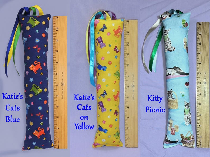 May include: Three cat toy pillows, each approximately 30 cm long, with colourful patterns and ribbons. One is navy blue with orange and green cats, one is yellow with cat and butterfly designs, and the last is light blue with cat illustrations. Text on the pillows reads "Katie's Cats Blue", "Katie's Cats on Yellow", and "Kitty Picnic".