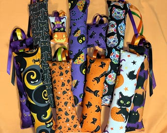 May include: A collection of rectangular fabric catnip toys featuring various Halloween-themed patterns including black cats, pumpkins, stars, and sugar skulls. Each toy is topped with a colorful ribbon loop for hanging, showcasing vibrant orange, purple, black, and white fabric prints.