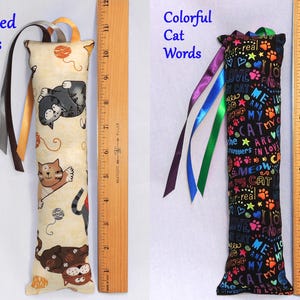 May include: Two cat-themed catnip toys. The first toy is tan with a pattern of cats and yarn balls. The second toy is black with colorful text that says "Me and My Cat are in Love". Both toys have ribbons attached.