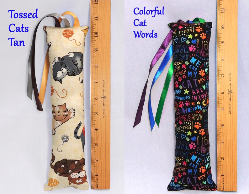 May include: Two cat-themed catnip toys. The first toy is tan with a pattern of cats and yarn balls. The second toy is black with colorful text that says "Me and My Cat are in Love". Both toys have ribbons attached.